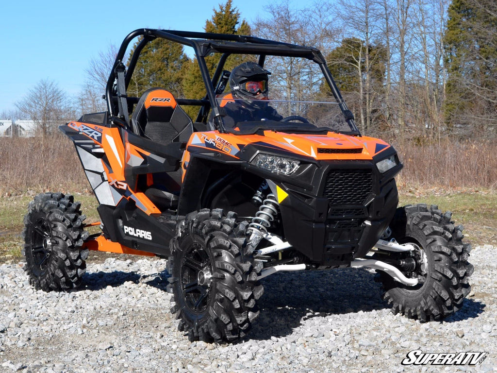 A Polaris RZR S 1000 fitted with a half windshield is parked on gravel. The helmeted driver sits in the open cab, surrounded by leafless trees and sparse grass beneath a clear sky.