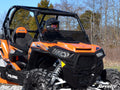 A Polaris RZR 900 equipped with a Half Windshield is parked on rocky terrain. A driver in a black helmet sits inside, surrounded by sparse trees beneath a clear sky. SuperATV appears in the lower right corner.
