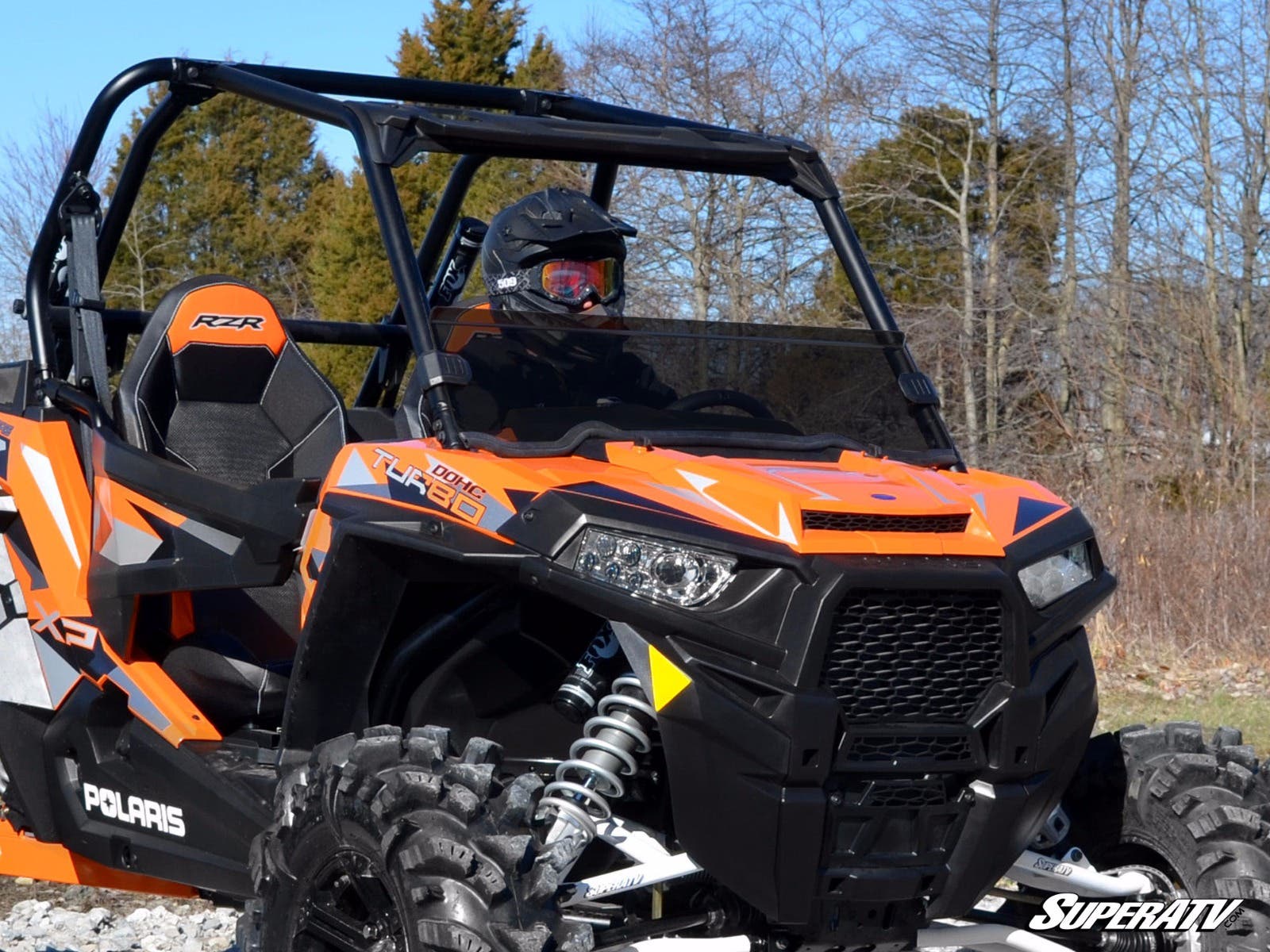 A Polaris RZR 900 equipped with a Half Windshield is parked on rocky terrain. A driver in a black helmet sits inside, surrounded by sparse trees beneath a clear sky. SuperATV appears in the lower right corner.