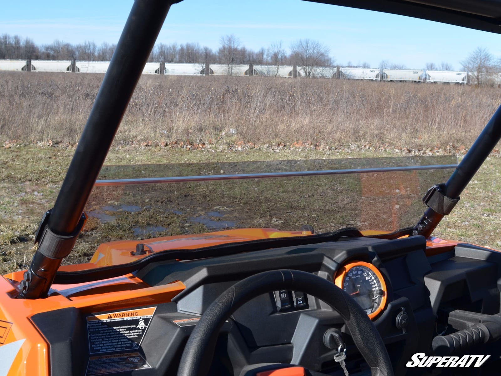 View from inside an orange off-road vehicle featuring the Polaris RZR XP Turbo Half Windshield, dashboard, and steering wheel. Parked on grass with mud ahead, white railcars and trees are visible under a clear blue sky. SuperATV logo shown.