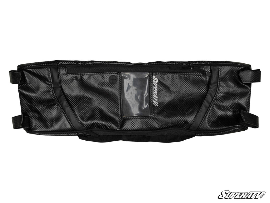 The Polaris RZR 900 Overhead Bag is black and rectangular with a carbon fiber texture, multiple pockets, a clear central phone holder, zipper closures, end mounting straps, and the SuperATV logo on the bottom right.