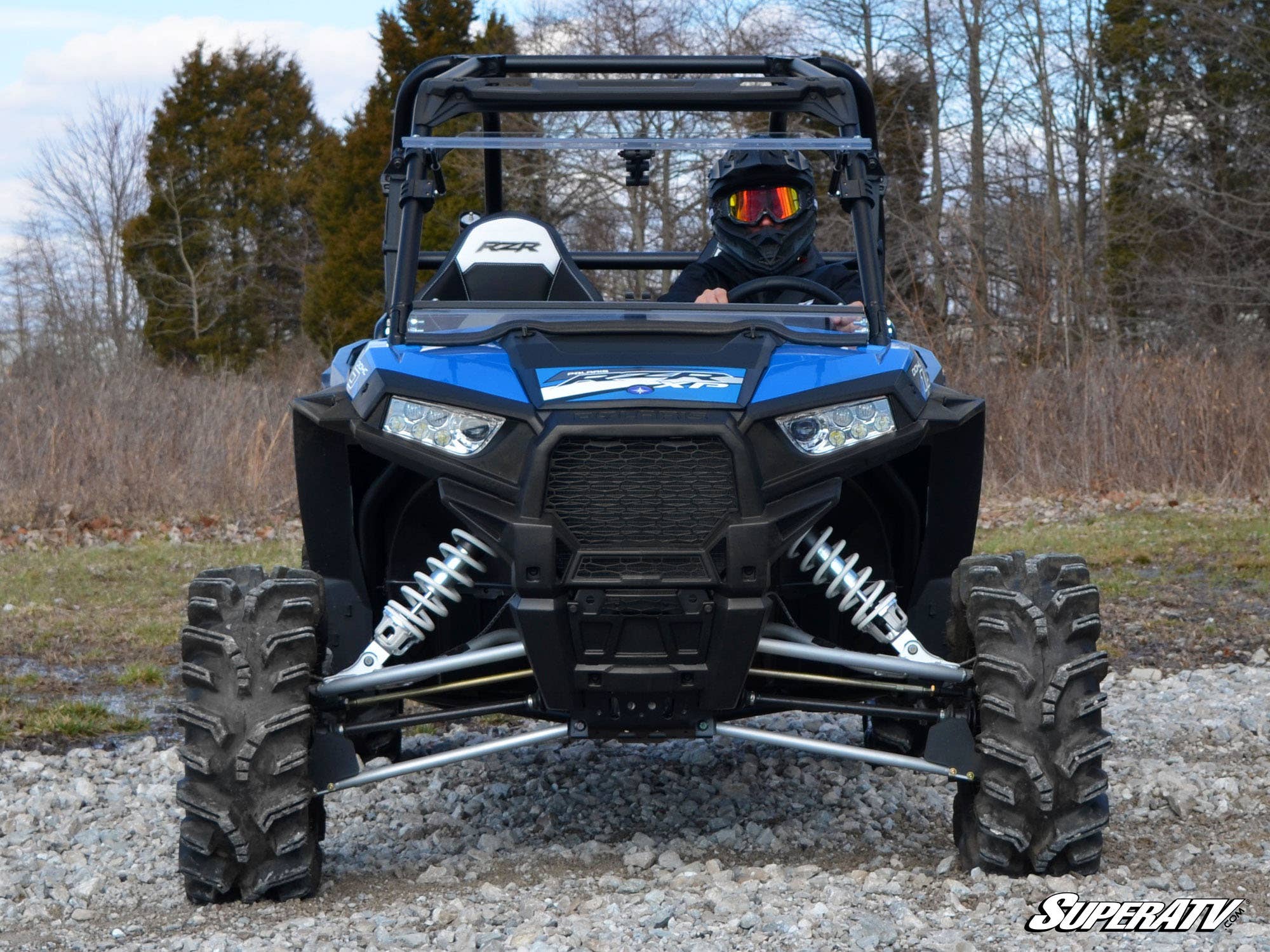 A helmeted driver pilots a blue Polaris RZR equipped with a Polaris RZR XP Turbo Flip Windshield and large, rugged tires over rocky terrain. Leafless trees and clouds are in the background, with the SuperATV logo at the bottom right.