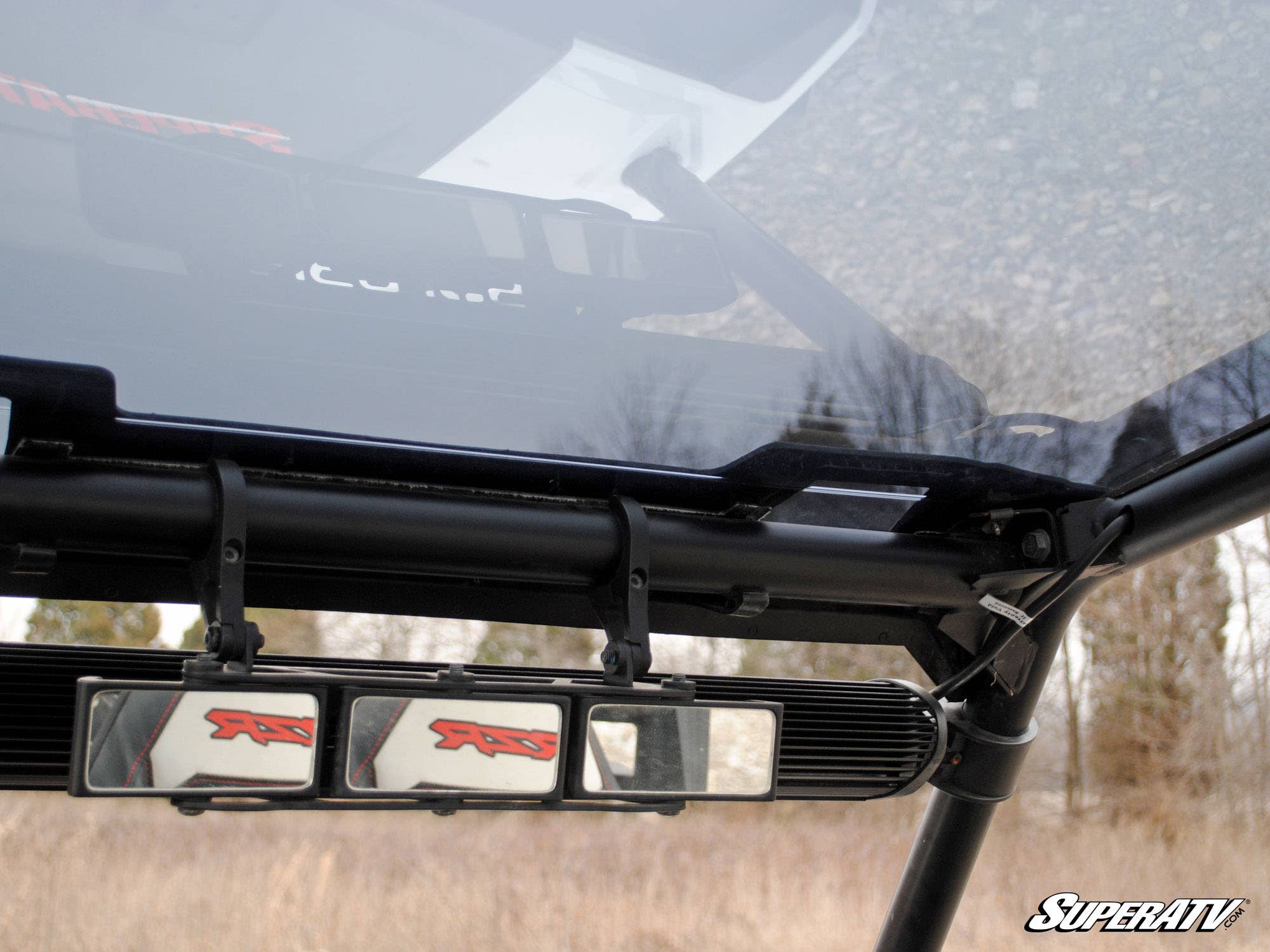 Close-up of a Polaris RZR XP Turbo Tinted Roof with a rearview mirror assembly featuring three panels, each bearing the red RZR logo. Dry grass, bare trees, and a cloudy sky fill the background. SuperATV is visible in the lower right corner.
