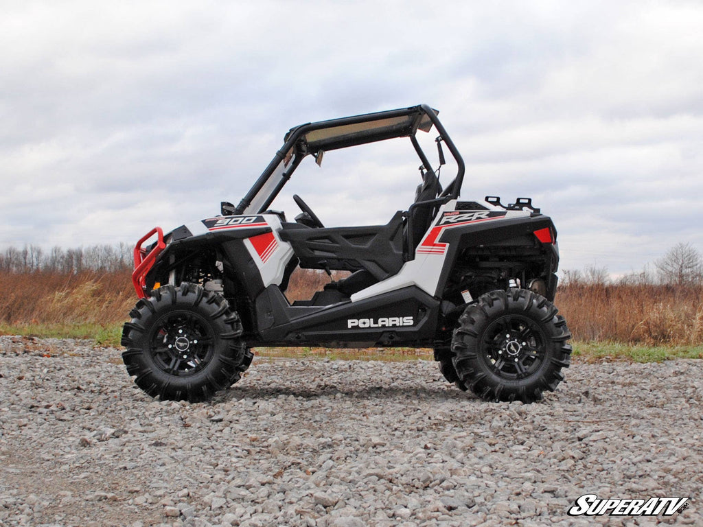 A Polaris RZR 900 with a 2 Lift Kit sits on rocky ground outdoors under a cloudy sky, surrounded by leafless trees and brown grass. It features large off-road tires, a roll cage, and SuperATV branding in the bottom right corner.