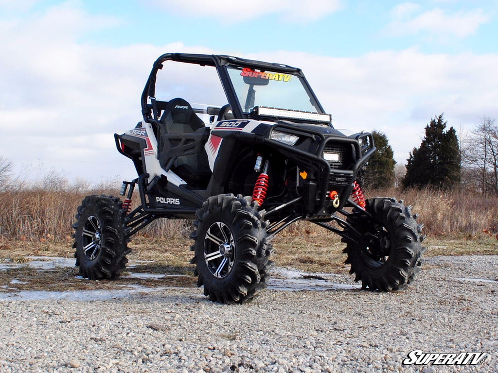 A black and white Polaris ATV with a RZR S 1000 7-10 Lift Kit, large rugged tires, red suspension springs, and an open two-seat cabin is parked on rocky ground outdoors. Sparse trees and a cloudy sky are in the background.
