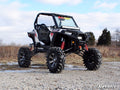 A black and white Polaris ATV with a RZR S 1000 7-10 Lift Kit, large rugged tires, red suspension springs, and an open two-seat cabin is parked on rocky ground outdoors. Sparse trees and a cloudy sky are in the background.