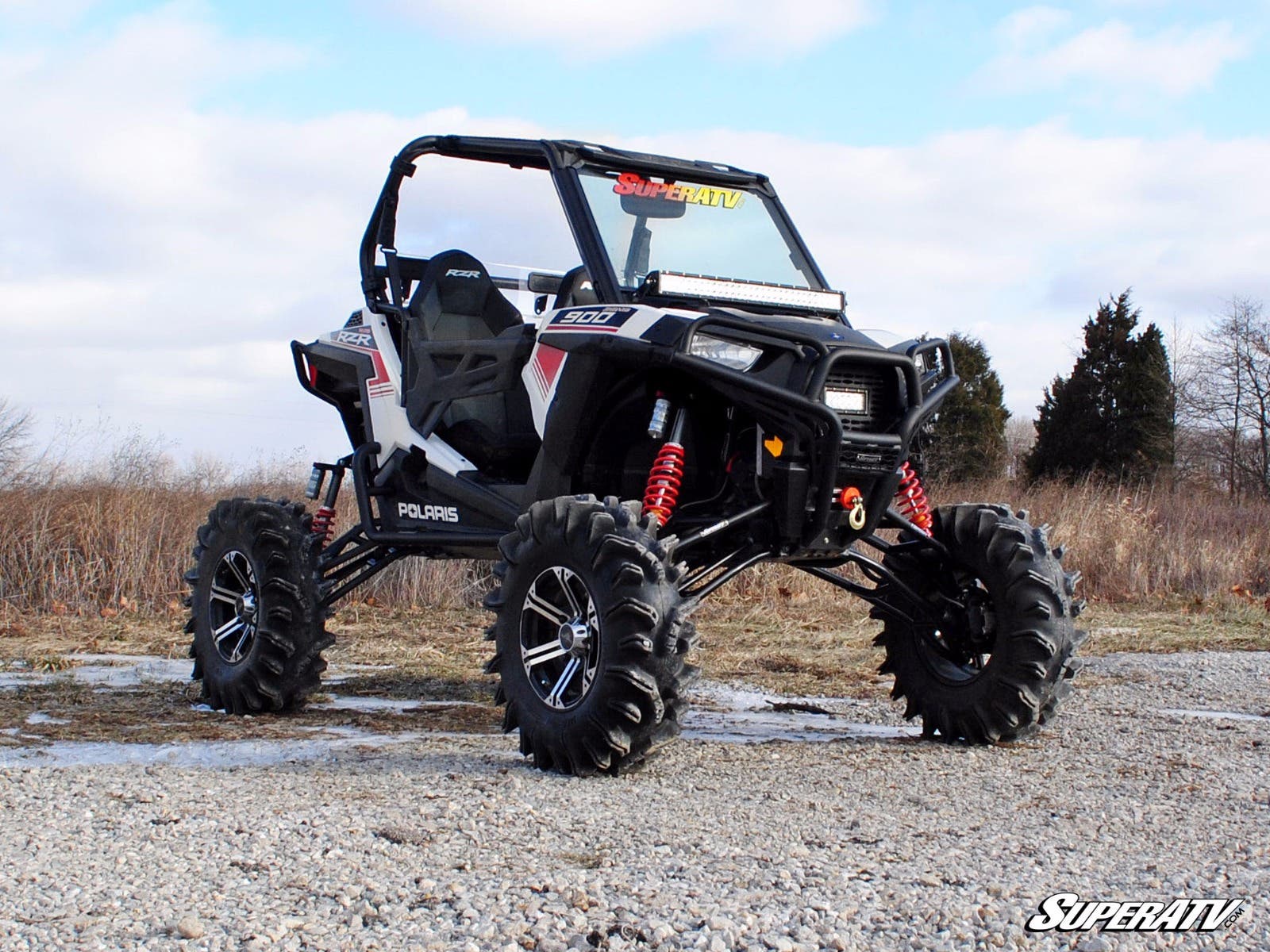 A black and white Polaris ATV with a RZR S 1000 7-10 Lift Kit, large rugged tires, red suspension springs, and an open two-seat cabin is parked on rocky ground outdoors. Sparse trees and a cloudy sky are in the background.