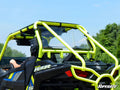 A Polaris RZR S 1000 with a bright yellow roll cage and black body, featuring a SuperATV rear windshield, is driven outdoors on a sunny day with green trees and a blue sky in the background.