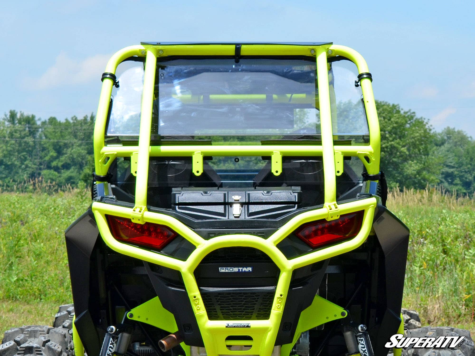 A Polaris RZR 900 with a rear windshield is shown from the back on grass with trees behind it. The off-road vehicle features red tail lights, black panels, and a SuperATV logo, highlighting its rugged, outdoor-ready style.