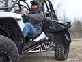 A person in a helmet and sunglasses sits in a white off-road vehicle with an open door, large tires, and metal frame bars on rocky terrain. Polaris RZR Door Bags are featured. Bare trees and a cloudy sky are visible. SuperATV is shown bottom right.