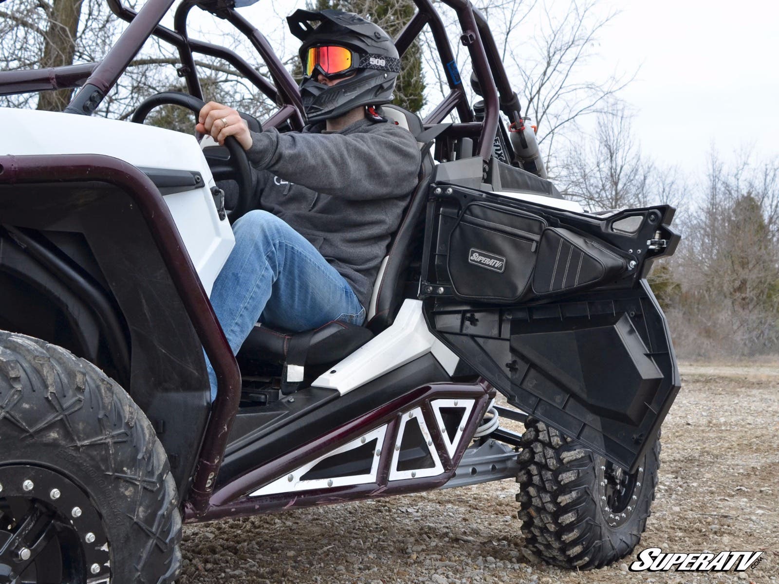 A person in a helmet and sunglasses sits in a white off-road vehicle with an open door, large tires, and metal frame bars on rocky terrain. Polaris RZR Door Bags are featured. Bare trees and a cloudy sky are visible. SuperATV is shown bottom right.