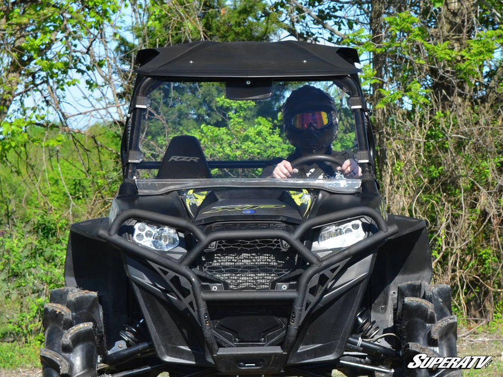 A person wearing a helmet and goggles sits in a black off-road ATV equipped with a Polaris RZR Full Windshield, featuring rugged tires and a front grill guard. Lush green foliage and trees are in the background, with the SuperATV logo at the bottom right.