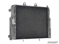 The Polaris RZR Heavy-Duty Radiator features a metal build with horizontal cooling fins, two top mounting tabs, side inlet and outlet pipes, a rugged surface, and the SuperATV logo in the lower right corner.
