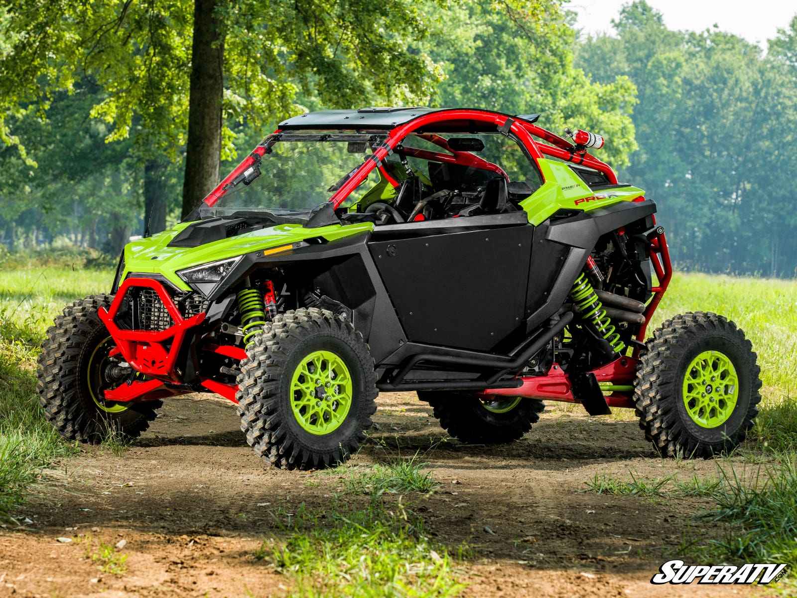 A Polaris RZR PRO XP with SuperATV Aluminum Doors is parked on a dirt trail in the woods, showing off its large rugged tires, visible suspension, roll cage, and black aluminum doors. Sunlight filters through the trees onto this off-road vehicle.