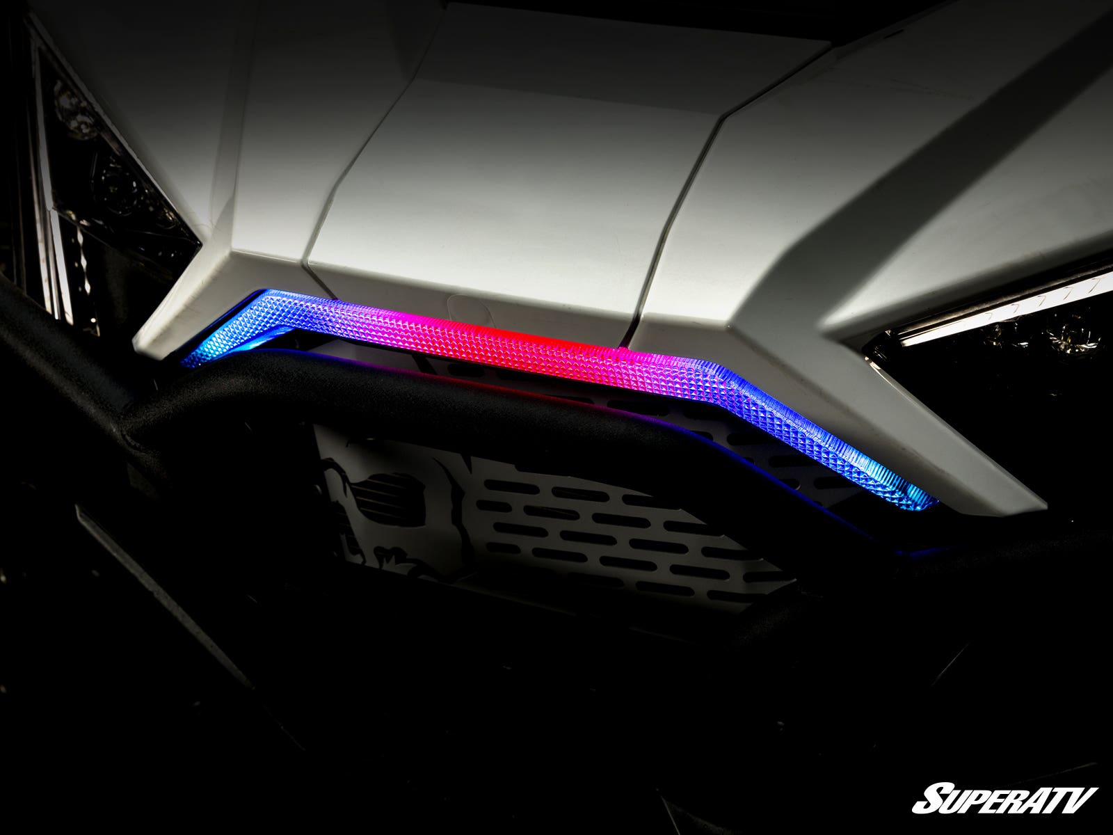 Close-up of a Polaris RZR Pro R Front Accent Light on a white vehicle’s angular hood, featuring a red center LED and blue side LEDs. The SuperATV logo appears in the bottom right, with illuminated accents standing out in the dark image.