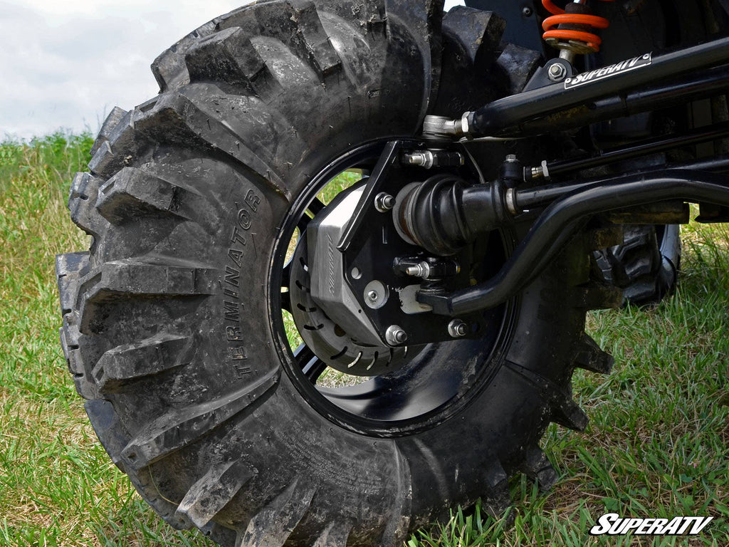 Close-up of a rugged off-road tire with deep treads on grass, revealing the suspension, axle, and brake components installed with the Polaris RZR 800 4 Portal Gear Lift. The SuperATV logo is visible in the bottom right corner.