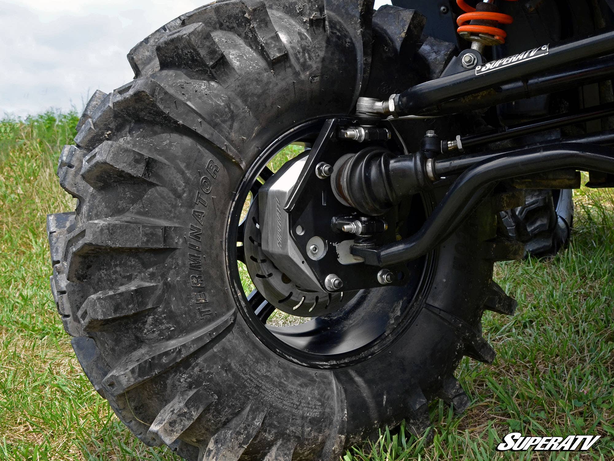 Close-up of a rugged off-road tire with deep treads on grass, revealing the suspension, axle, and brake components installed with the Polaris RZR 800 4 Portal Gear Lift. The SuperATV logo is visible in the bottom right corner.