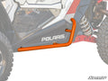 Polaris RZR XP 1000 Heavy-Duty Nerf Bars in orange are mounted along the lower side panel, protecting the body near the front and rear wheels. These nerf bars run parallel to the ground above rugged tires and a visible suspension coil.