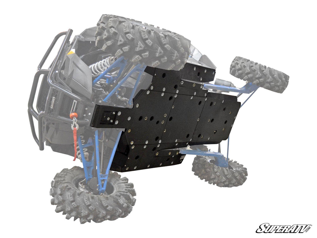 A Polaris RZR XP 1000, caked in mud, is tilted to reveal a large black reinforced skid plate covering the underside. Its suspension stands out against a faded background, emphasizing the Polaris RZR XP 1000 Full Skid Plate. SuperATV logo appears bottom ri