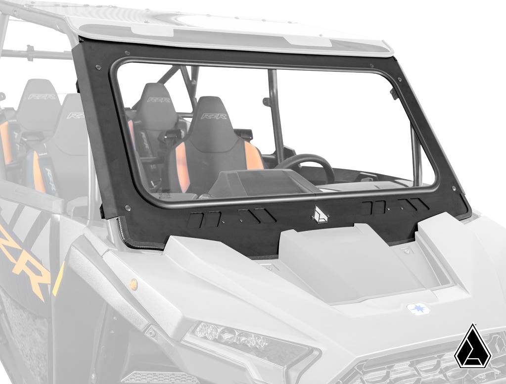 A close-up of the Assault Industries Polaris RZR XP Glass Windshield installed on a gray off-road utility vehicle with two front seats and part of a steering wheel visible. The clear glass shows the interior, and a small triangular logo is on the lower fr