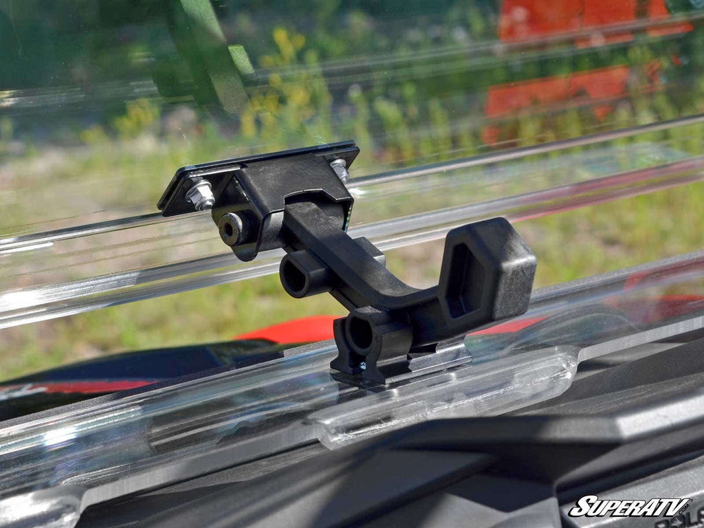 Close-up of a black windshield clamp latch securing a clear Polaris RZR S 1000 Flip Windshield on a vehicle. The background shows blurred greenery and red, with SuperATV branding in the lower right corner.