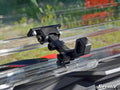 Close-up of a black windshield clamp latch securing a clear Polaris RZR S 1000 Flip Windshield on a vehicle. The background shows blurred greenery and red, with SuperATV branding in the lower right corner.