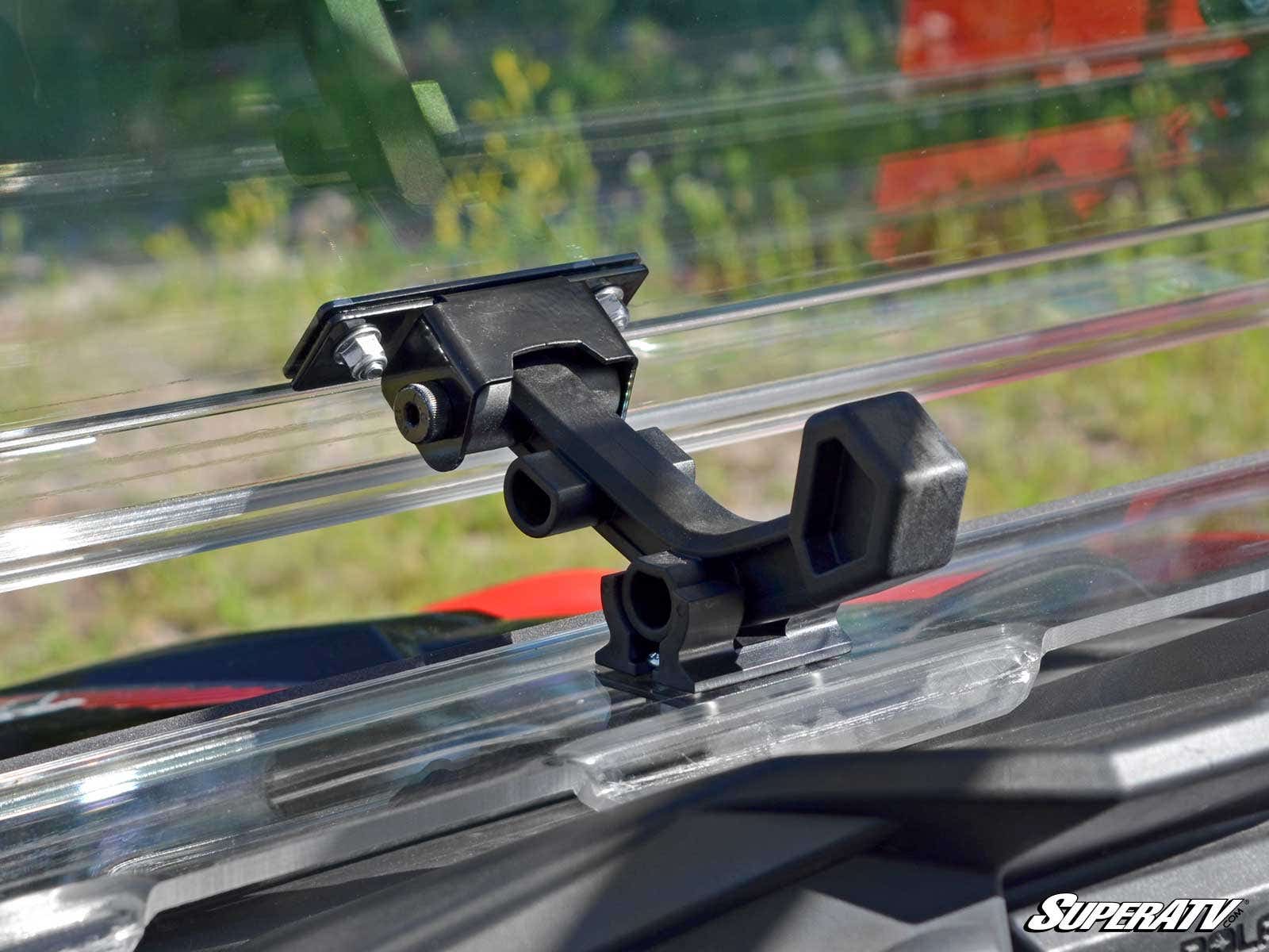 Close-up of a black windshield clamp latch securing a clear Polaris RZR S 1000 Flip Windshield on a vehicle. The background shows blurred greenery and red, with SuperATV branding in the lower right corner.