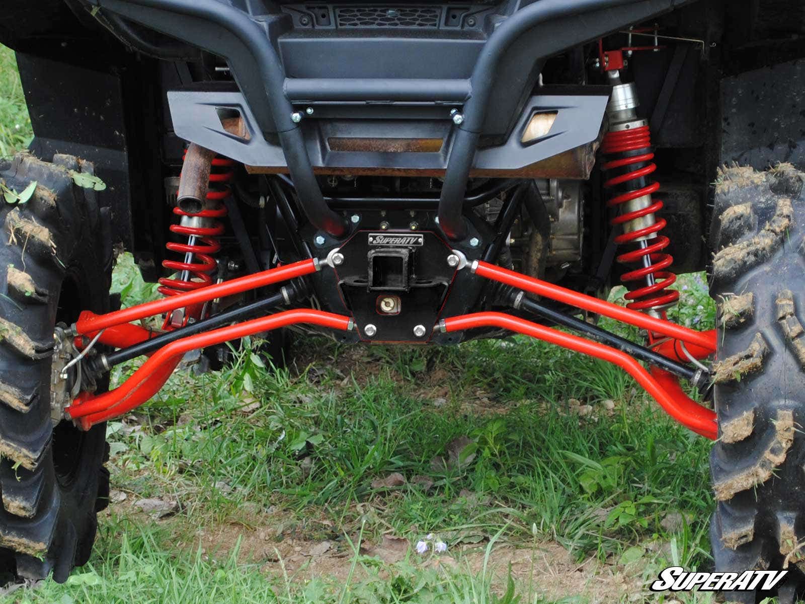 Close-up of a Polaris RZR XP 900 Rear Receiver Hitch installed, featuring rugged rear suspension, bright red shock absorbers, mud-treaded off-road tires on green grass. The SuperATV logo is in the bottom right corner.