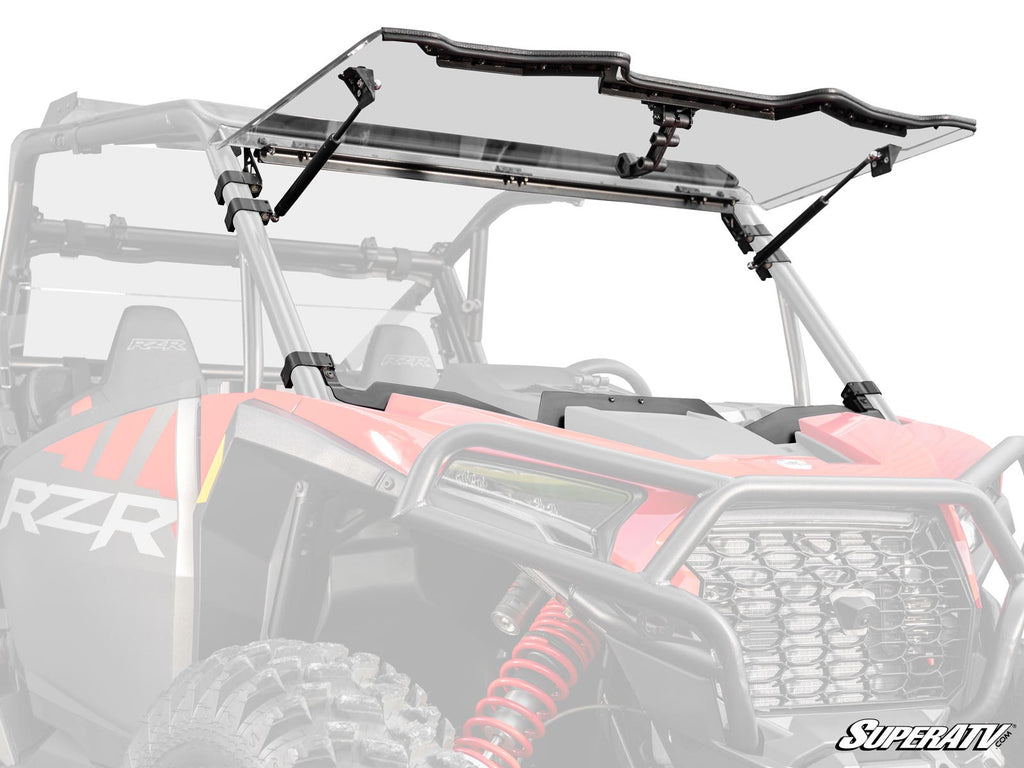 A Polaris RZR XP with a clear, flip-up windshield from SuperATV is shown in red. The background and parts of the vehicle are faded to highlight the Polaris RZR XP Flip Windshield and its mounting brackets, with the SuperATV logo in the lower right corner.