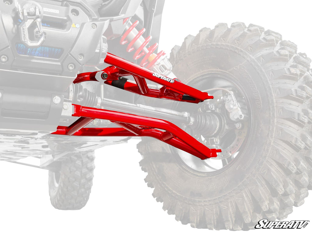 A close-up of glossy, red Polaris RZR XP High-Clearance A-Arms installed on an off-road vehicle. The detailed suspension is attached to a rugged tire, with the faded vehicle in the background and the SuperATV logo in the bottom right corner.