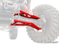 A close-up of glossy, red Polaris RZR XP High-Clearance A-Arms installed on an off-road vehicle. The detailed suspension is attached to a rugged tire, with the faded vehicle in the background and the SuperATV logo in the bottom right corner.