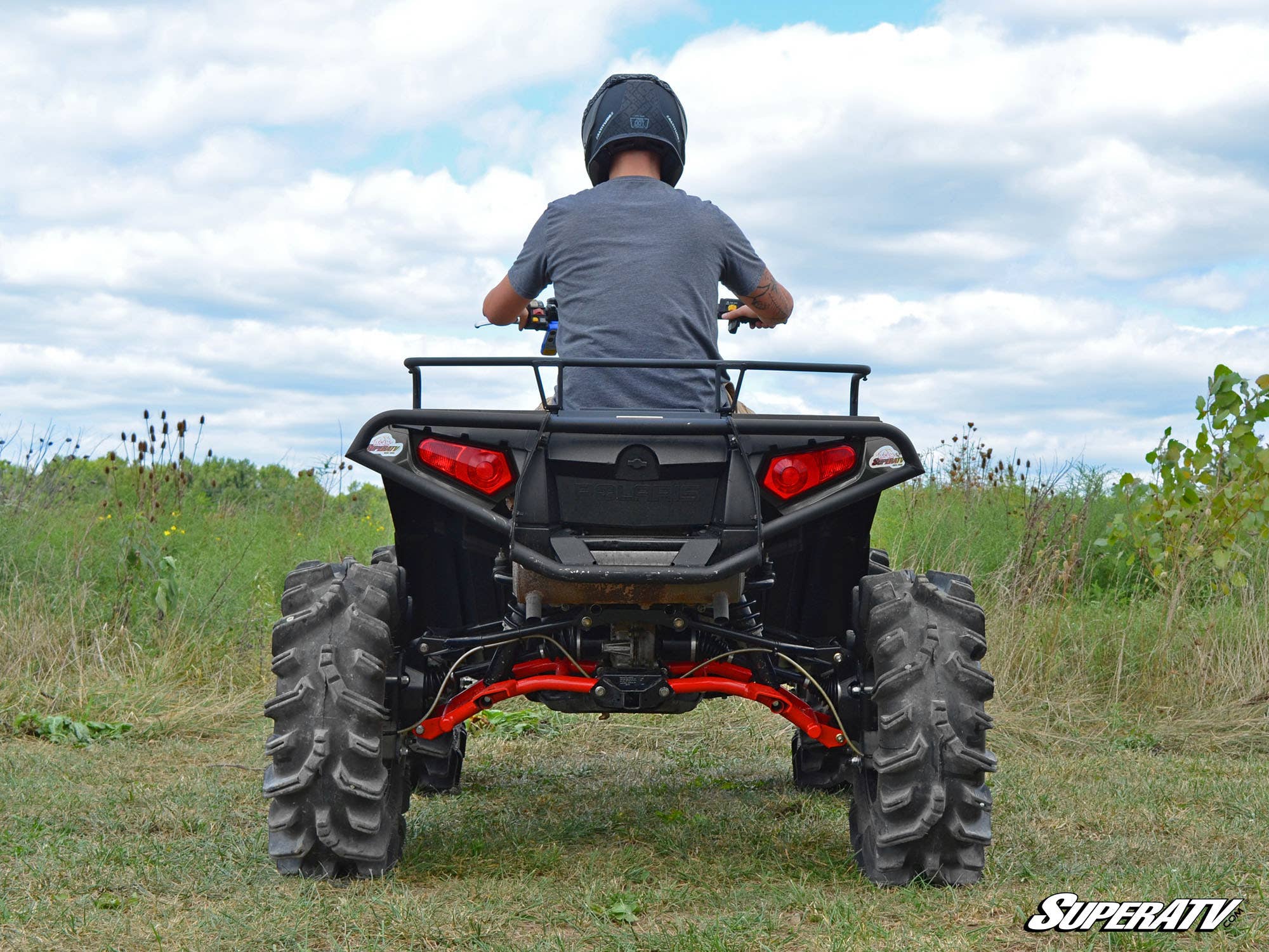 A person in a helmet and gray shirt sits on a black ATV with large rugged tires, red suspension, and rear cargo rack, featuring the Polaris Sportsman XP 4 Portal Gear Lift, in an open grassy field under a partly cloudy sky.