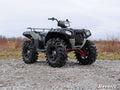 A Polaris Scrambler with a 2 Lift Kit and large mud tires is parked on rocky gravel in a grassy field under clouds. The ATV features a front rack, raised suspension, and “SuperATV” branding visible on the image corner.