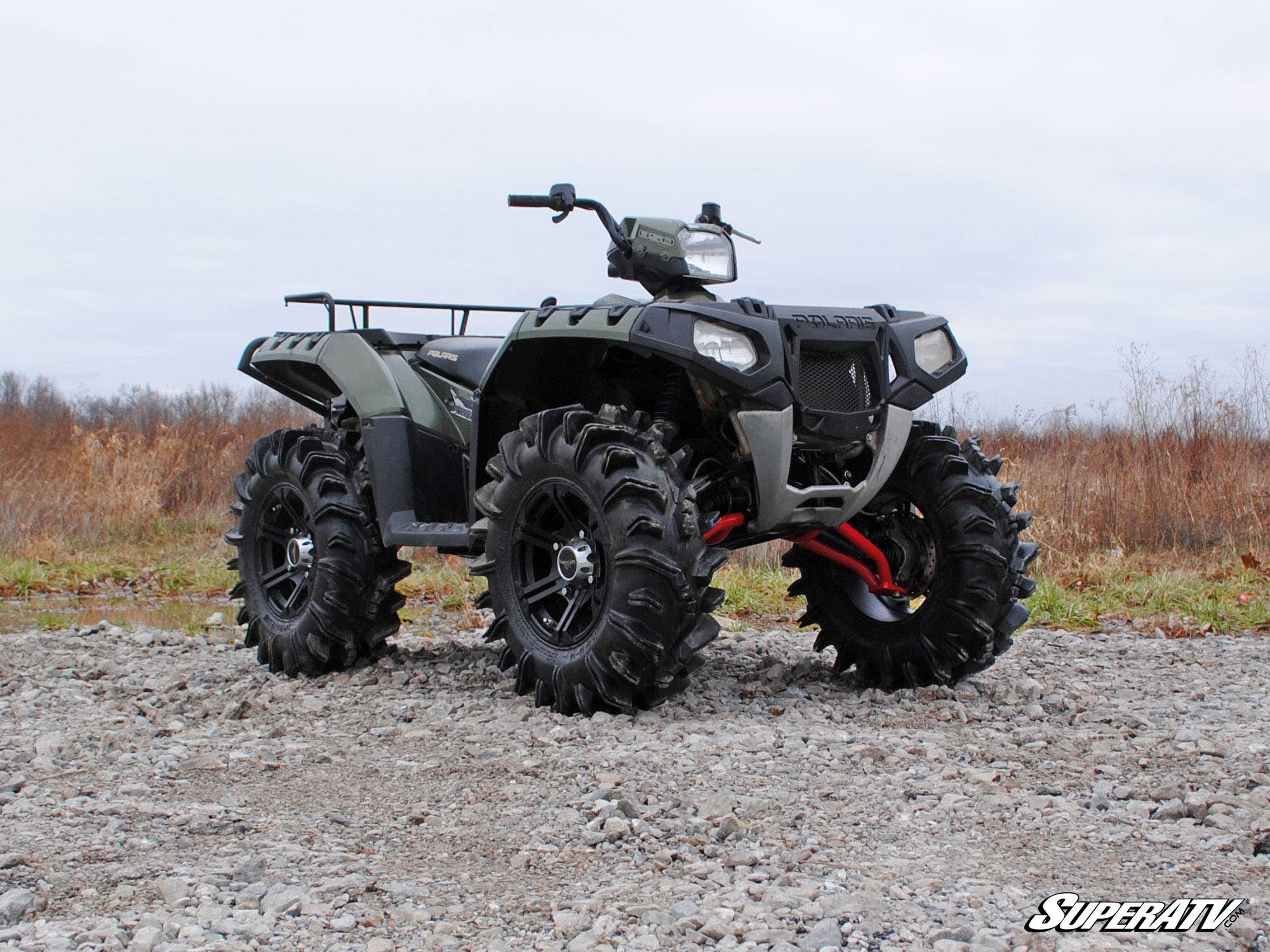 A Polaris Scrambler with a 2 Lift Kit and large mud tires is parked on rocky gravel in a grassy field under clouds. The ATV features a front rack, raised suspension, and “SuperATV” branding visible on the image corner.