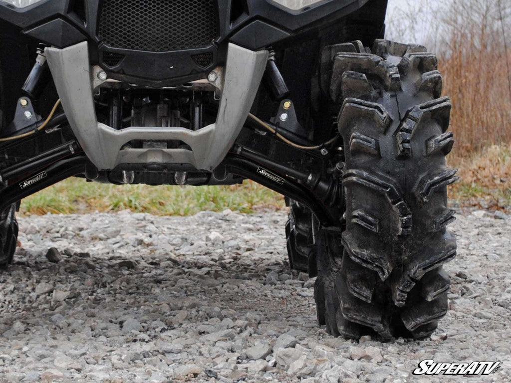 Close-up of the front of an off-road ATV equipped with Polaris Sportsman XP High Clearance A-Arms, featuring aggressive mud tires and robust suspension. The ATV is parked on rocky ground with dry grass and bare trees. SuperATV logo visible bottom right.