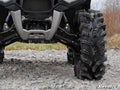 Close-up of the front of an off-road ATV equipped with Polaris Sportsman XP High Clearance A-Arms, featuring aggressive mud tires and robust suspension. The ATV is parked on rocky ground with dry grass and bare trees. SuperATV logo visible bottom right.