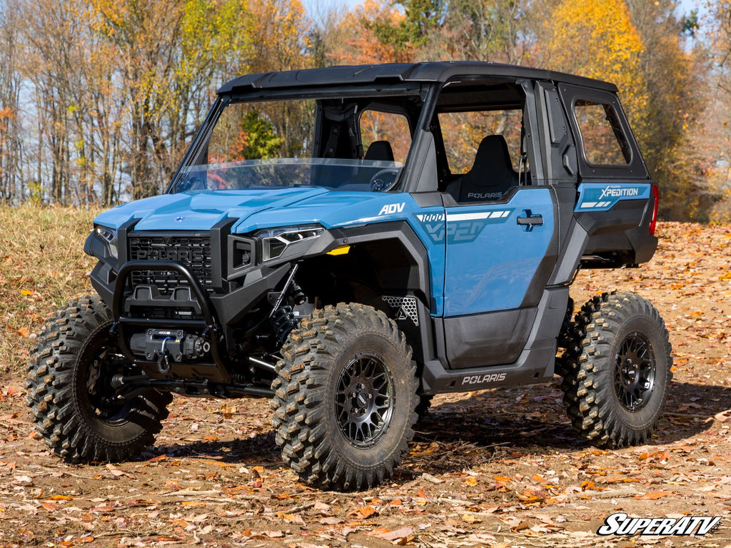 A blue Polaris Xpedition fitted with a Half Windshield is parked on an autumn leaf-covered path, framed by orange and yellow trees under a clear sky. The SuperATV logo appears in the bottom right corner.