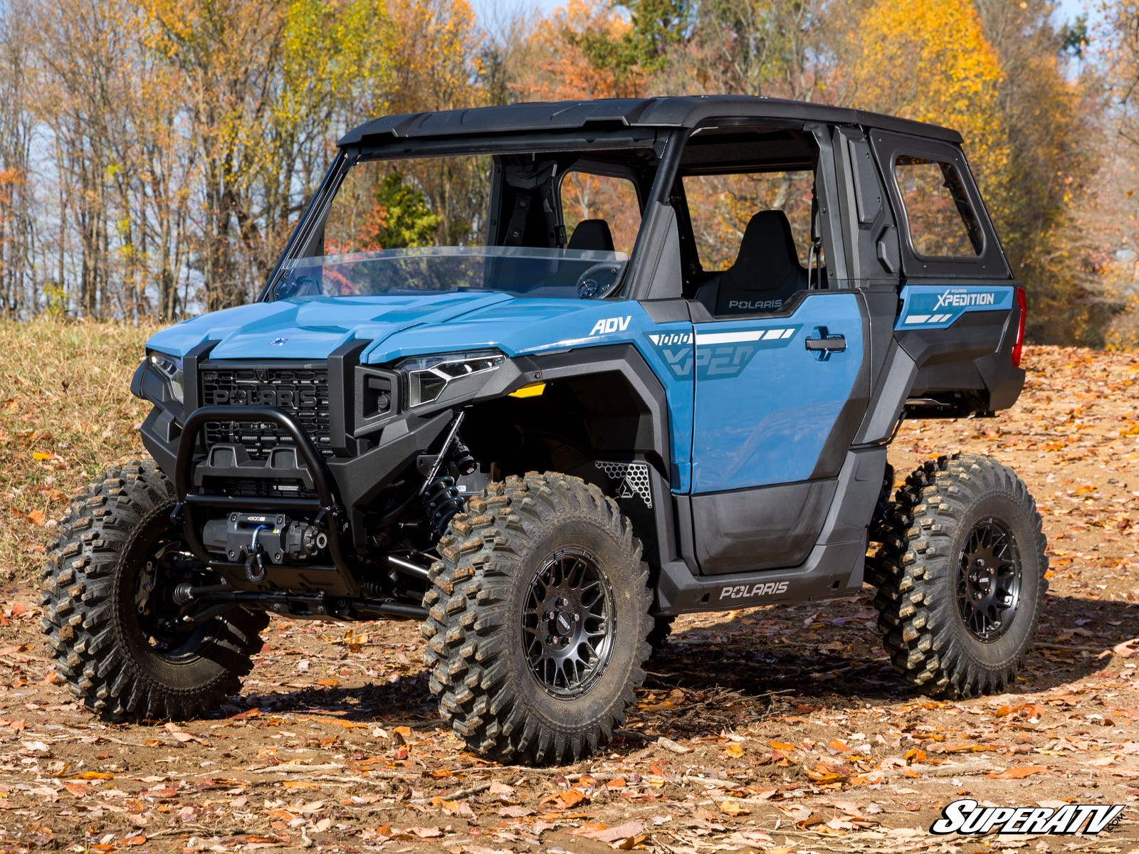 A blue Polaris Xpedition fitted with a Half Windshield is parked on an autumn leaf-covered path, framed by orange and yellow trees under a clear sky. The SuperATV logo appears in the bottom right corner.