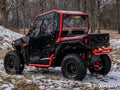 A Polaris General XP with Scratch Resistant Full Cab Doors is parked on snowy forest ground, featuring black paint, red accents, rugged tires, enclosed clear windows, a rear cargo area, and SuperATV branding on the vehicle and corner.