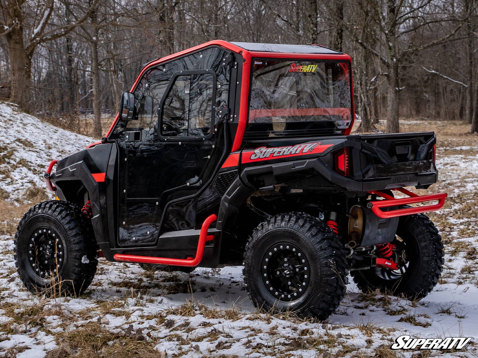 A Polaris General XP with Scratch Resistant Full Cab Doors is parked on snowy forest ground, featuring black paint, red accents, rugged tires, enclosed clear windows, a rear cargo area, and SuperATV branding on the vehicle and corner.