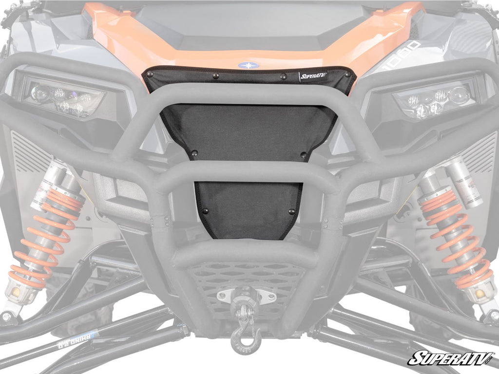 Close-up of a Polaris General Grille Cover featuring a black mesh design, orange suspension springs, sturdy metal bumper, headlights, and a winch hook at the bottom center. The SuperATV logo appears in the lower right corner.