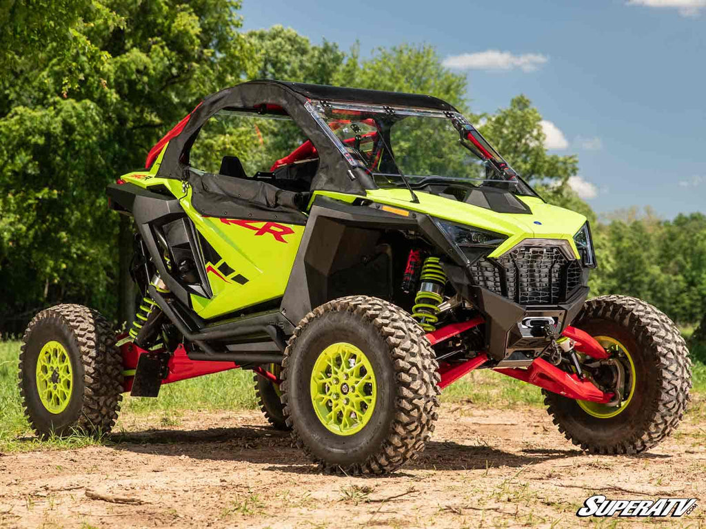 A Polaris RZR Pro S equipped with a Winch Mounting Plate sits on dirt. It features neon yellow and black colors, red suspension, large rugged tires, tinted windows, and a roll cage. Trees and blue sky are in the background. SuperATV logo is at bottom righ