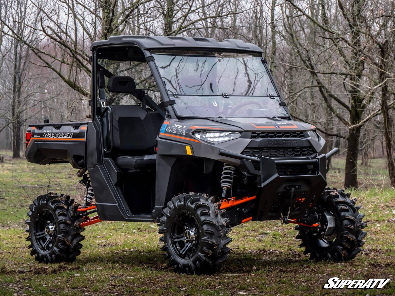 A black Polaris Ranger 1000 with large tires and orange suspension is parked in a forest clearing, featuring a Flip Down Windshield cab and doors. The SuperATV logo appears in the bottom right corner.