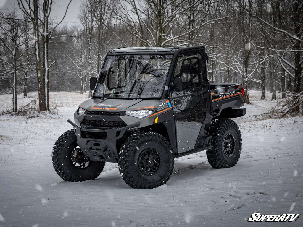 A Polaris Ranger XP 1000 equipped with High-Clearance 2 Forward A-Arms features large off-road tires and is parked on snowy ground in a wintry forest. Snow falls gently, leafless trees surround it, and the SuperATV logo appears at bottom right.