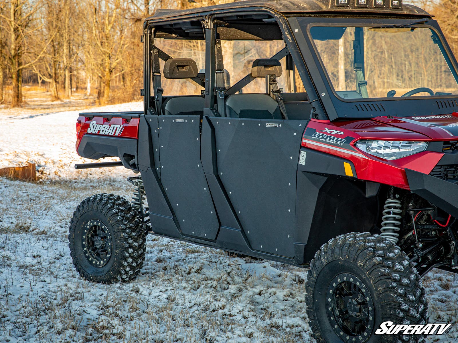 A Polaris Ranger 1000 with black Aluminum Doors by SuperATV, rugged tires, and red body is parked on a snowy grassy area near leafless trees under a clear sky.
