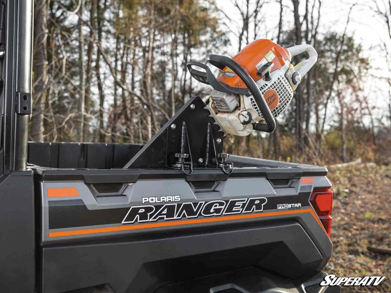 A Polaris Ranger Chainsaw Mount secures a chainsaw in the cargo bed of a Polaris Ranger ProStar. Leafless trees and grass in the background suggest an outdoor setting in late fall or early spring.