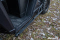 Close-up of the lower side of a Polaris Ranger XP 1000 with Heavy-Duty Nerf Bars, featuring black metal bars and a textured step. Ground below shows grass, leaves, and some snow. The “SuperATV” logo appears in the bottom right corner.