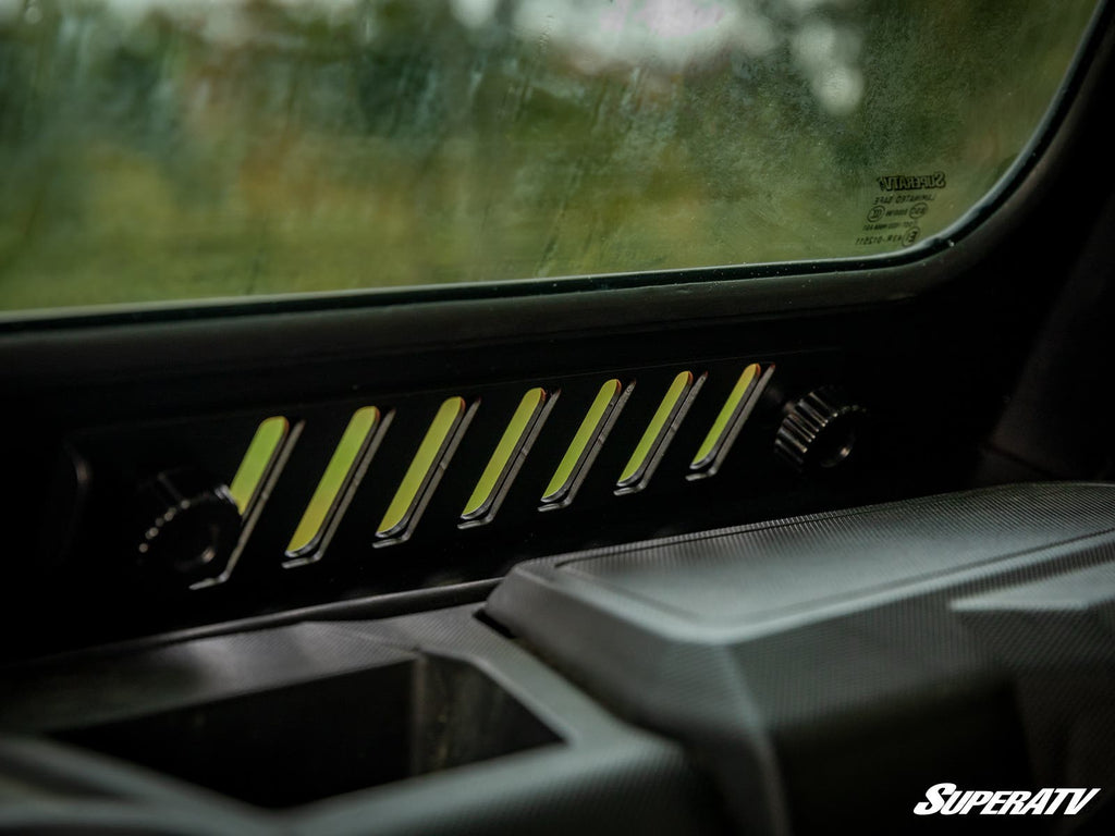 Close-up of a Ranger XP 1000 Glass Windshield interior panel featuring six angled vents, two round control dials on each side, the SuperATV logo in the lower right corner, and blurred greenery visible through the slightly dirty glass.