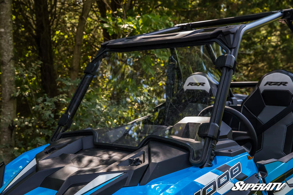 A close-up of a Polaris RZR XP Turbo with a full windshield and two black seats labeled RZR is parked outdoors among green leafy trees, sunlight filtering through the branches and illuminating the scene.