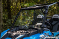 A close-up of a Polaris RZR XP Turbo with a full windshield and two black seats labeled RZR is parked outdoors among green leafy trees, sunlight filtering through the branches and illuminating the scene.